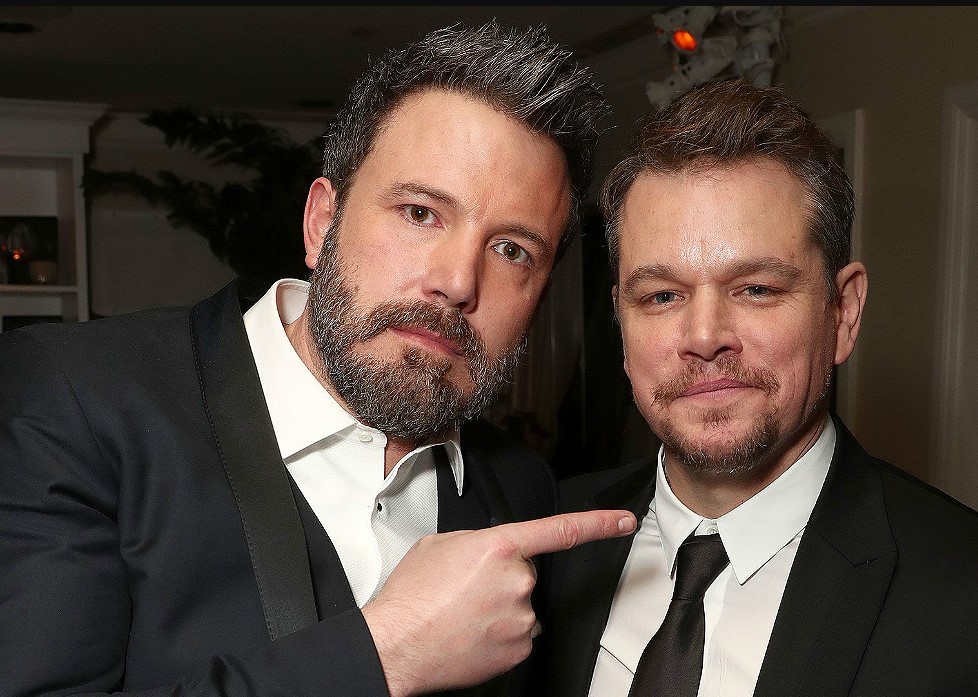 imagem em close do Ben Affleck a esquerda e Mat Dammon a direita posando para uma foto. Os dois estão de terno preto 