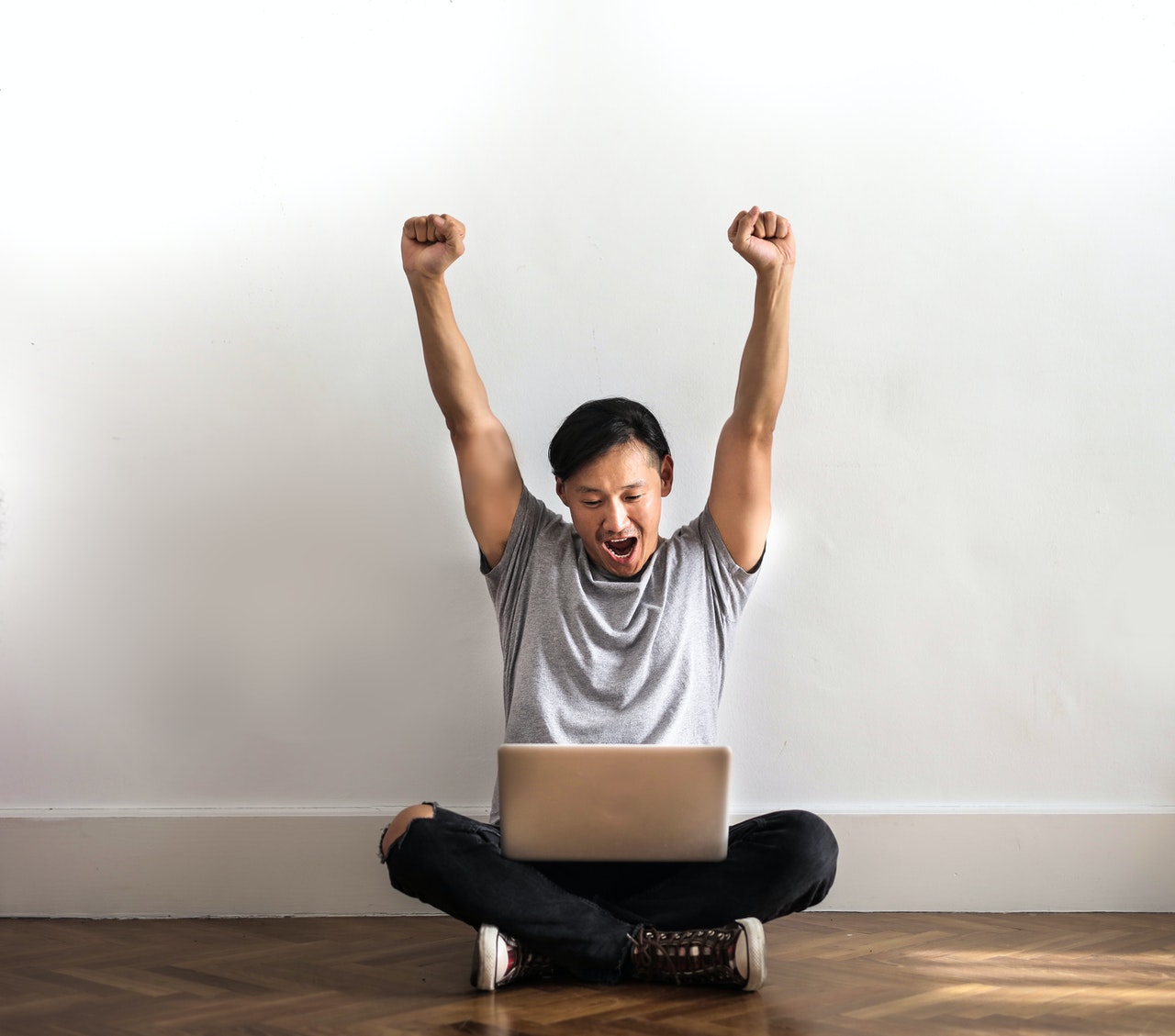 homem de camiseta cinza e calça preta comemora sentado ao chão com seu laptop no colo