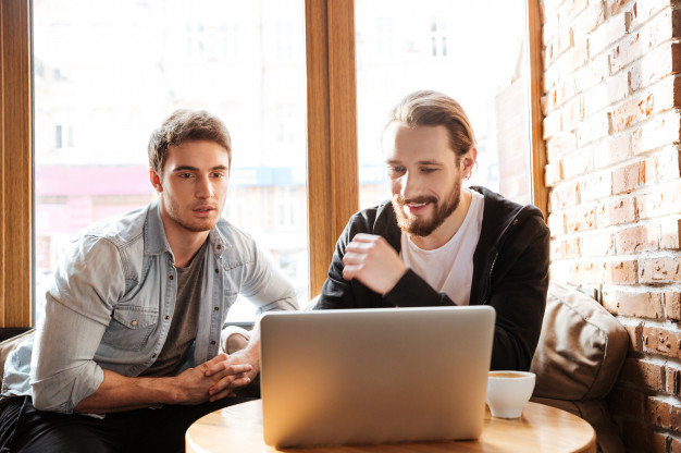 dois homens usando camisa jeans e outro com casaco preto sorriem de frente para computador em cima de uma mesa de um estabelecimento comercial