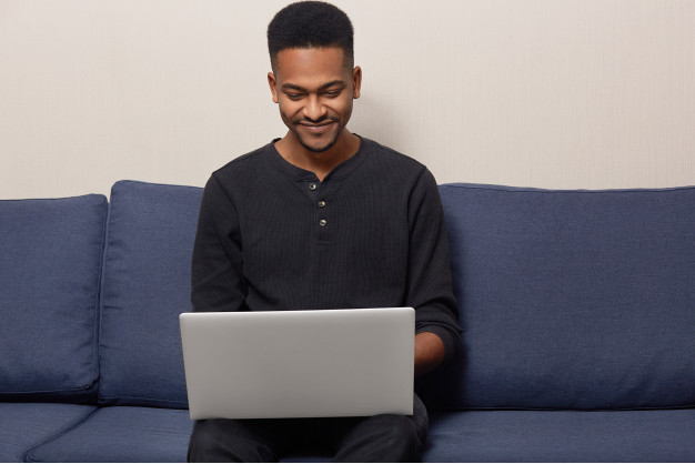 homem sentado em sofá azul usando roupas pretas sorrindo com um laptop prateado no colo