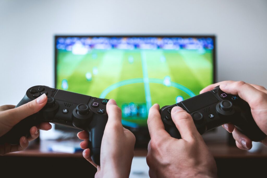 Mãos segurando joystick de videogame diante de uma tela de televisão com o jogo Fifa