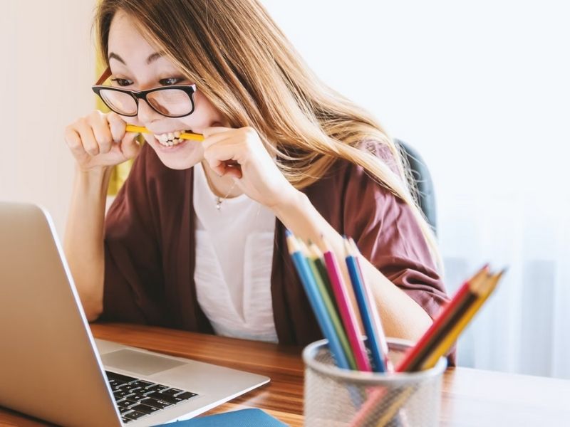 mulher em frente a um notebook mordendo um lápis enquanto uma vasilha com vários lápis coloridos está ao lado do computador